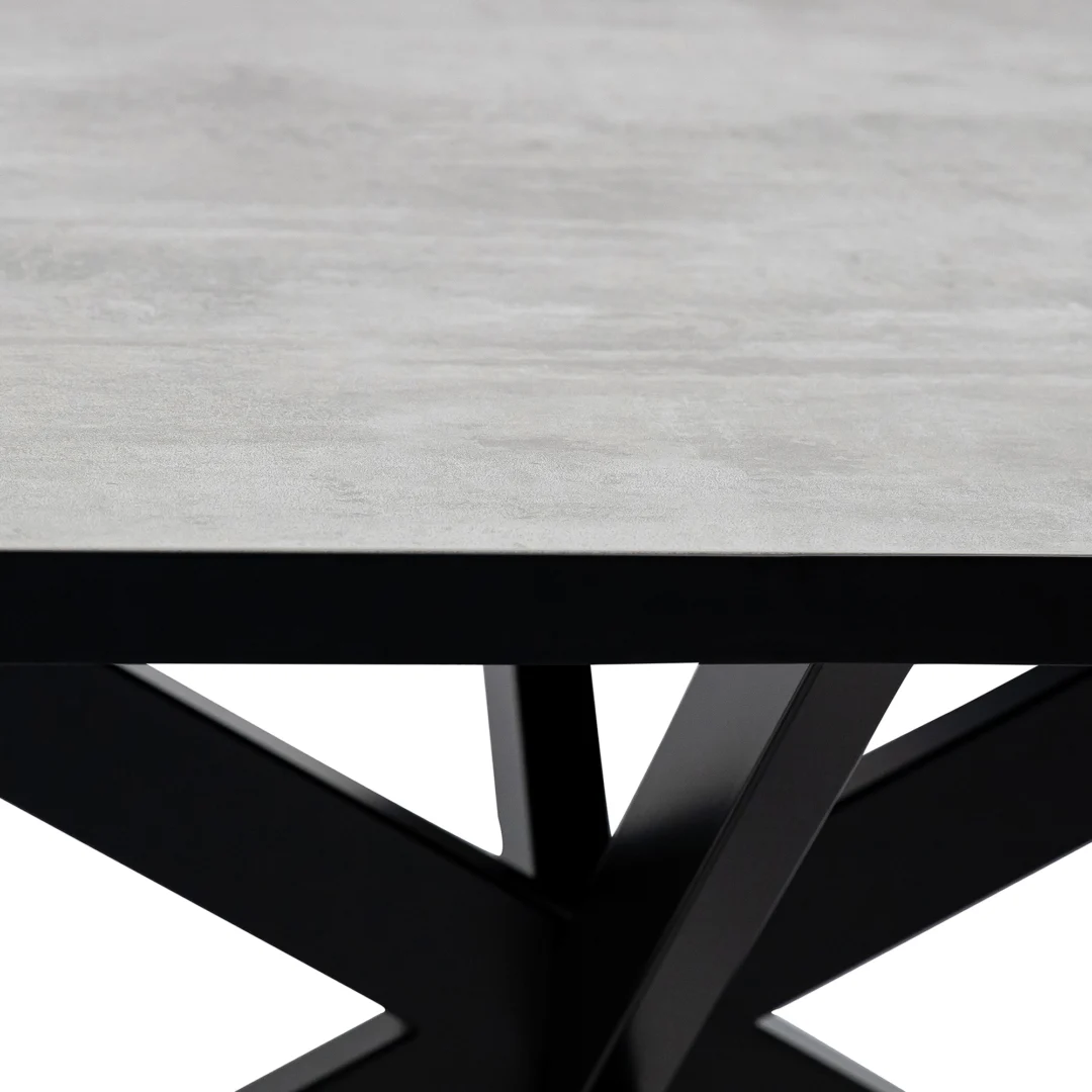 Een close-up van de Stalux Plat ovale eettafel 'Noud' 210 x 100 in zwart/beton, met een lichtgrijs, gestructureerd blad en zwarte metalen poten in een hoekig kriskras ontwerp.