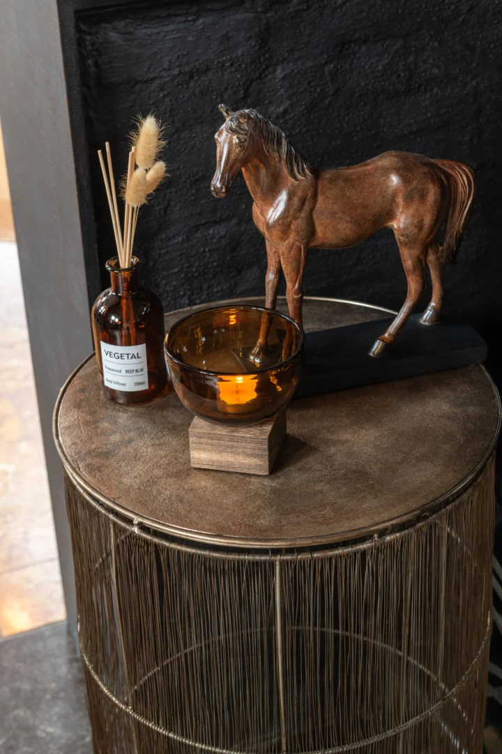 Een vierkant, bruin en goudkleurig aluminium bijzettafeltje (41 x 72 cm) bevat een bronzen paardenfiguurtje, een amberkleurige glazen kom met houten onderstel en een bruine fles met de naam Vegetal met gedroogd gras, tegen een donkere achtergrond.