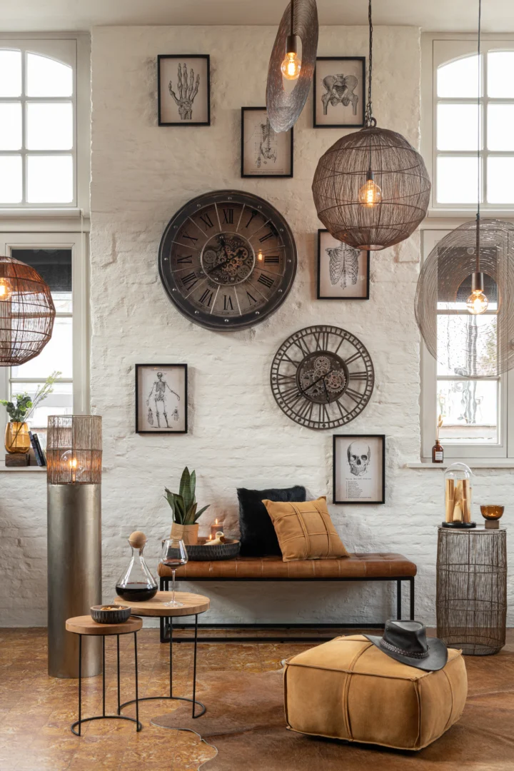 Een woonkamer in industriële stijl heeft een bruine, gouden Bijzettafel (41 x 72 cm, vierkant aluminium), stijlvolle inrichting, poef met hoed, grote wandklokken, ingelijste kunst, geweven hangers, planten, witte bakstenen muur en hoge ramen.