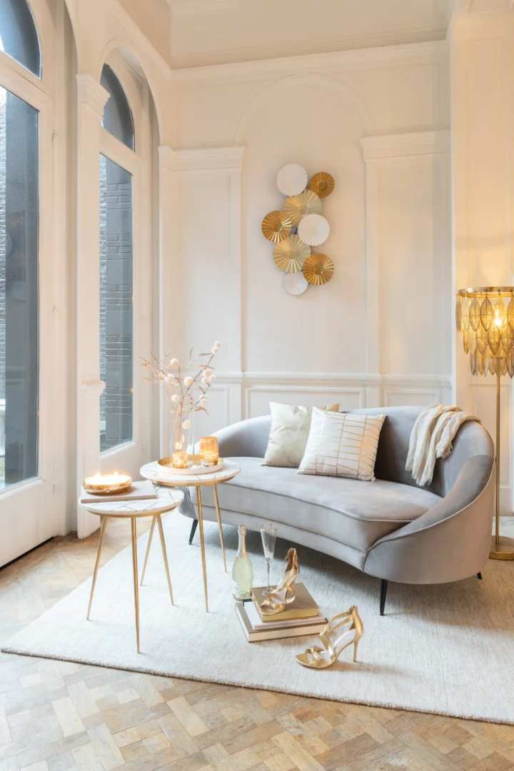 Een stijlvolle woonkamer met een gebogen grijze bank, neutrale kussens, een Bijzettafel Ivoor, Goud 41 x 46 CM Marmer versierd met kaarsen en decor, gouden hakken op boeken, een staande lamp, muurkunst en grote ramen die natuurlijk licht binnenlaten.