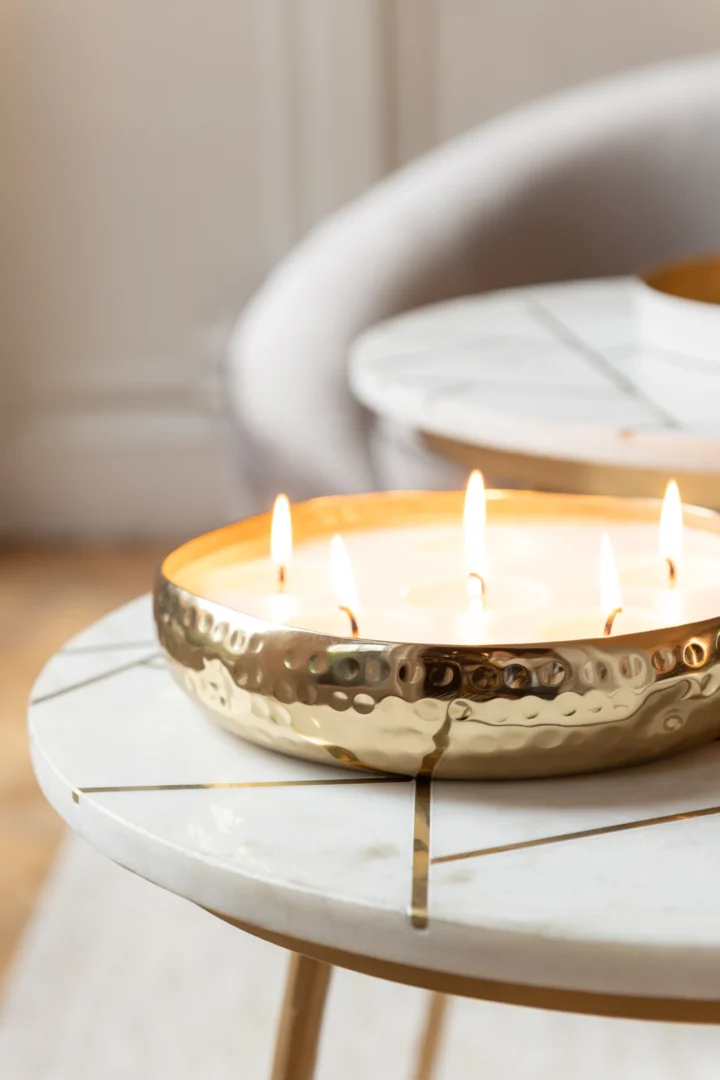 Een grote, gehamerd gouden metalen schaalkaars met meerdere vlammen staat op de Bijzettafel Ivoor, Goud 41 x 46 CM Marmer, een ronde wit marmeren tafel met gouden accenten. Op de achtergrond is een deel van een lichtgrijze stoel zichtbaar.