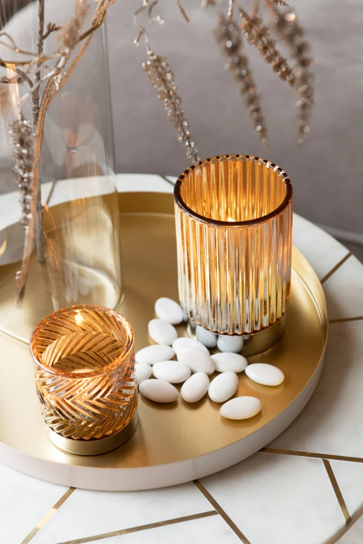 Op de Bijzettafel Ivoor, Goud 41 x 46 CM Marmer, toont een rond gouden dienblad twee verlichte glazen kaarsenhouders met patroon en witte ovale snoepjes, met decoratieve takken die erboven hangen.