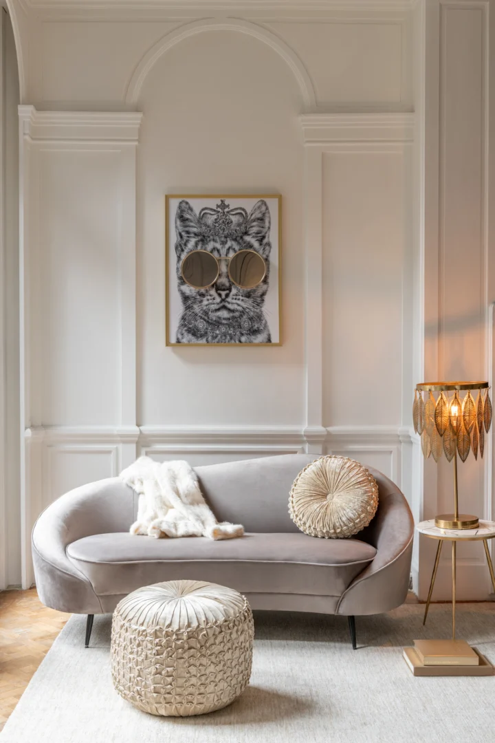 Een stijlvolle woonkamer met een gebogen grijze bank, een ronde voetenbank met structuur en de Bijzettafel Ivoor, Goud 41 x 46 CM Marmer als bijzettafel. Een ingelijste kattenillustratie met zonnebril hangt aan de witte wand boven de bank.
