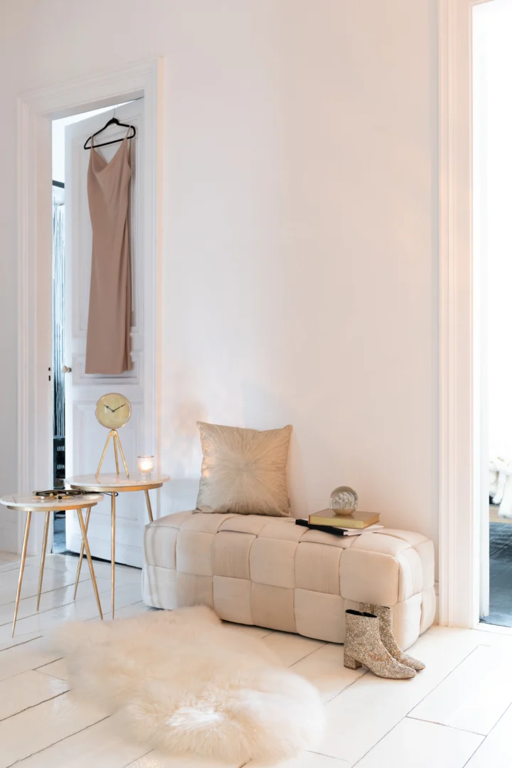 Een stijlvolle, minimalistische kamer met een beige poef, kussen, bontkleed en de Bijzettafel Ivoor Goud 41x46 CM Marmer met een klok, kaars en boeken. Op de vloer staan glinsterende laarzen en op de achtergrond hangt een bruine jurk aan de deur.