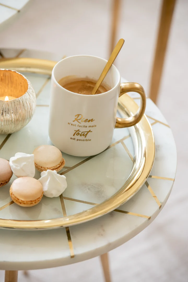 Op de Bijzettafel Ivoor, Goud 41 x 46 CM Marmer staat een kopje koffie met een gouden lepeltje, naast drie macarons en twee meringue koekjes. Op de mok staat Frans geschreven.