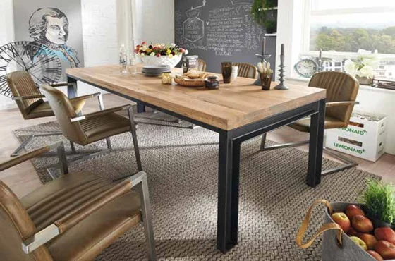 Een moderne eetkamer toont de Eettafel Herning Dessert - 180x100 tafel met metalen poten, omringd door bruine stoelen. De tafel is gedekt met borden, kaarsen en fruit, en er is een krijtbordmuur en grote ramen met een schilderachtig uitzicht.