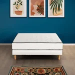 Witte boxspring 140x190 met houten poten op een vloerkleed. Drie ingelijste kunstwerken aan een blauwe muur. Heldere belichting.