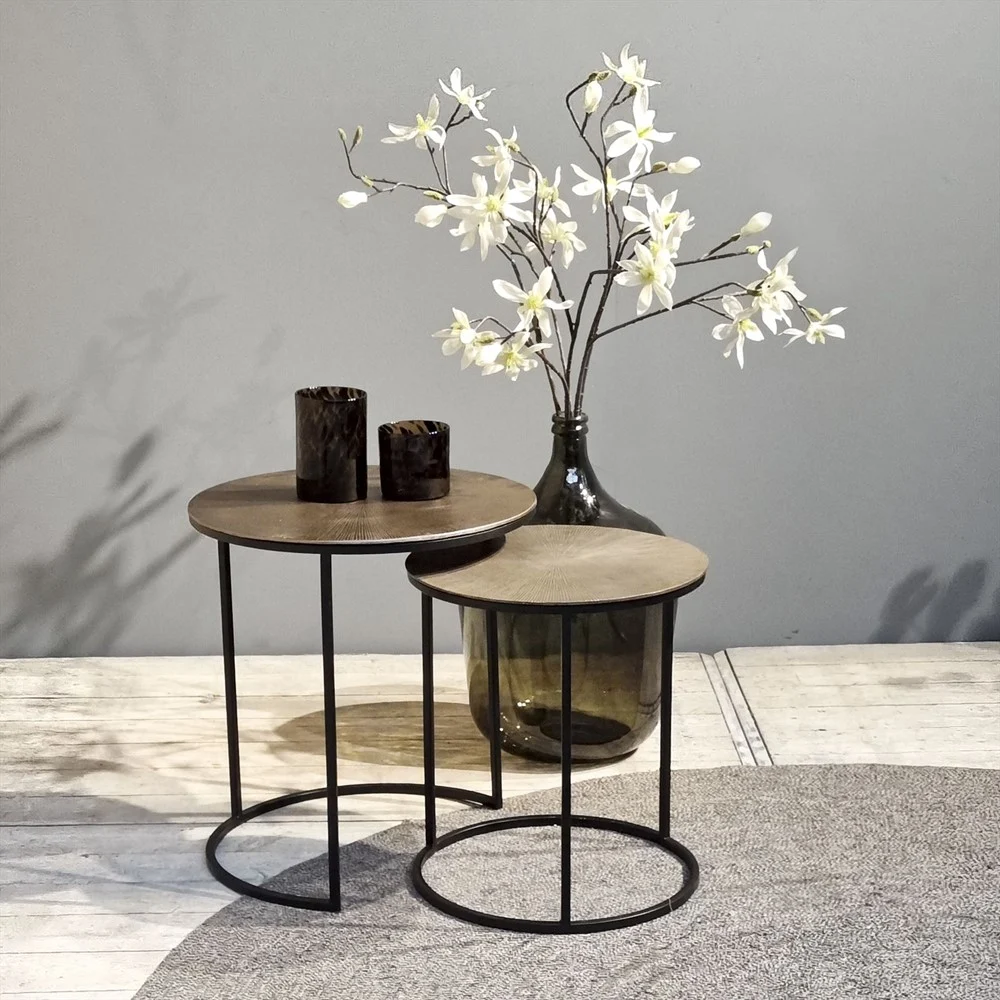 De Sidetable Ciara Set van 2 bestaat uit twee ronde nesttafels met metalen poten, met daarop een grote vaas van donker glas met witte kunstbloemen en twee donkere kandelaars, waardoor een minimalistische, neutrale setting ontstaat.
