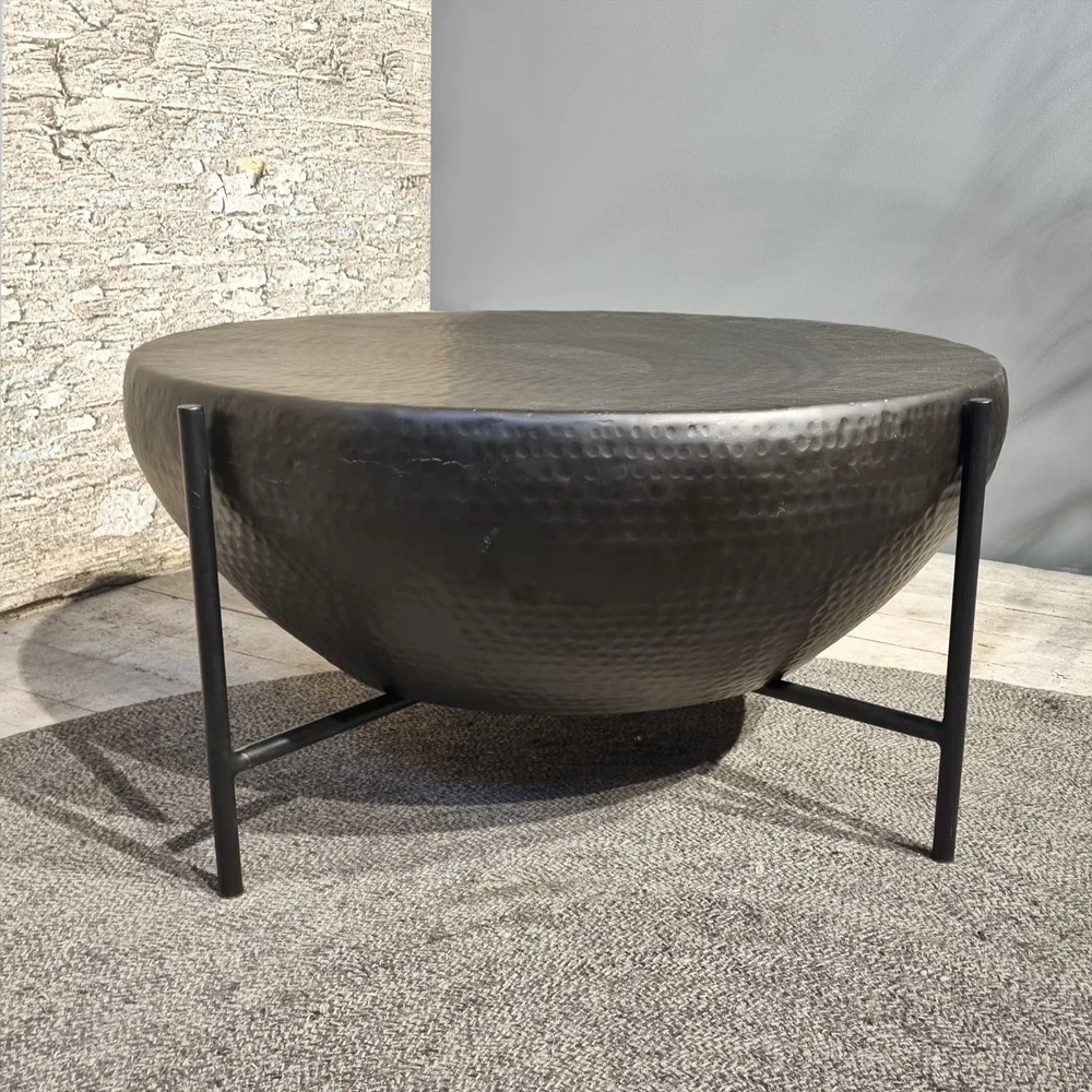 De Salontafel 85 x 85 CM Vierkant, een vierkante zwart gehamerde metalen salontafel met drie dunne zwarte poten, staat op een grijs tapijt tegen gestructureerde stenen en gladde grijze muren.