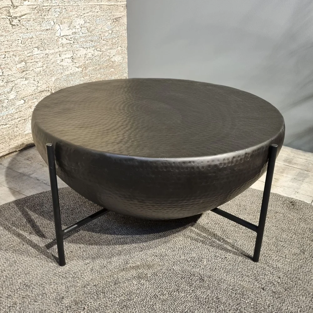 De Salontafel 85 x 85 CM Vierkant, met een donker gehamerd metalen blad en een zwart onderstel met drie poten, staat op een grijs vloerkleed met textuur voor een stenen en grijze muurachtergrond.
