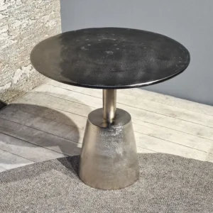 De Sidetable Kian Zwart Zilver, een klein rond zwart metalen tafeltje met een conische voet, staat op een lichthouten vloer bij een grijze muur en een stenen oppervlak met structuur, met een deel van een grijs vloerkleed in het zicht.