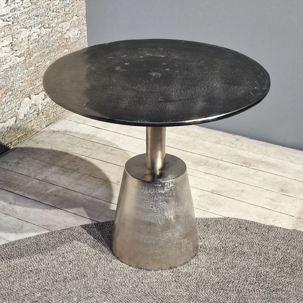 De Sidetable Kian Zwart Zilver, een klein rond zwart metalen tafeltje met een conische voet, staat op een lichthouten vloer bij een grijze muur en een stenen oppervlak met structuur, met een deel van een grijs vloerkleed in het zicht.