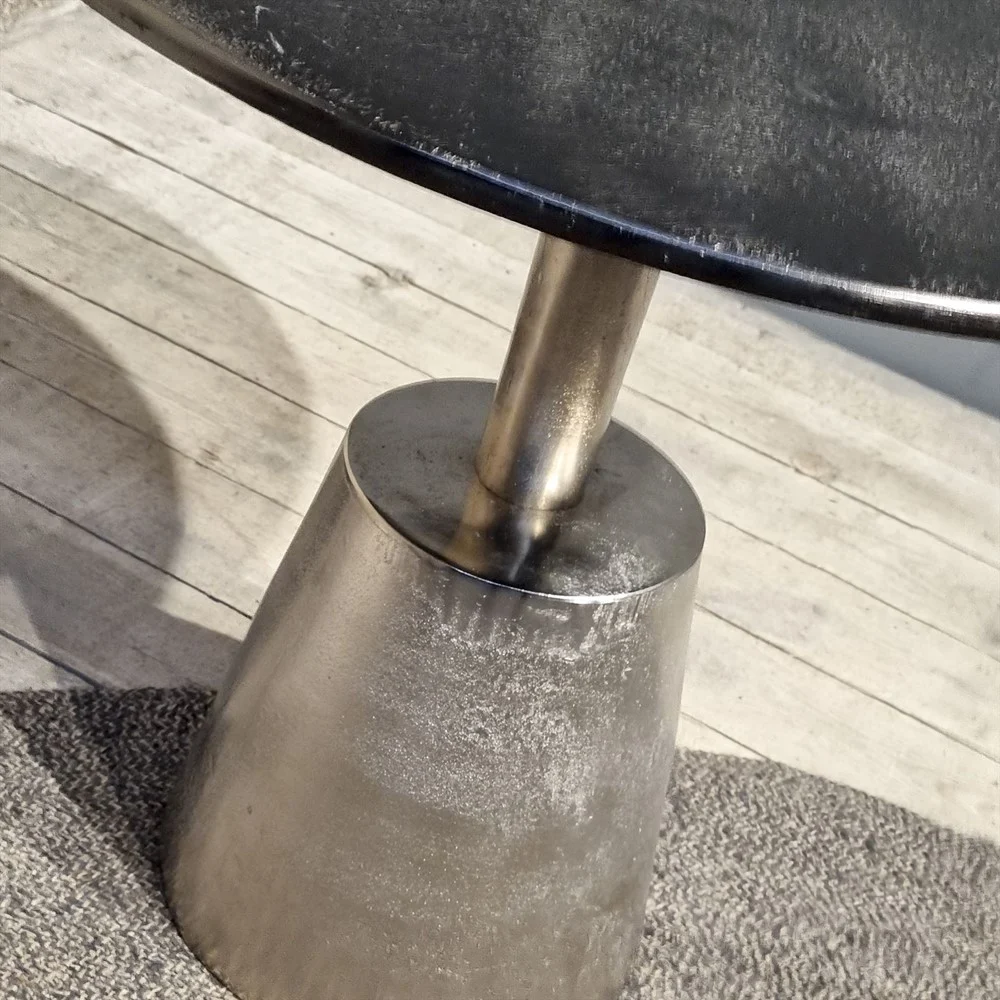 Een close-up van de Sidetable Kian Zwart Zilver, een klein rond metalen tafeltje met een glanzend kegelvormig onderstel, op een vloerkleed met textuur op houten planken.