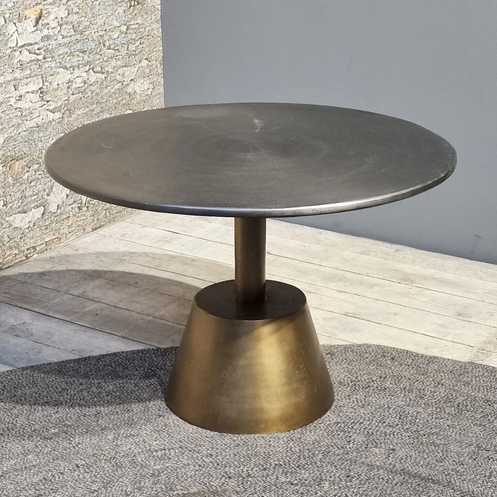 De Sidetable Kian Goud Zilver heeft een rond metalen blad en een stevig, kegelvormig bronzen onderstel, op een houten vloer naast een stenen muur met textuur en een grijs vloerkleed.