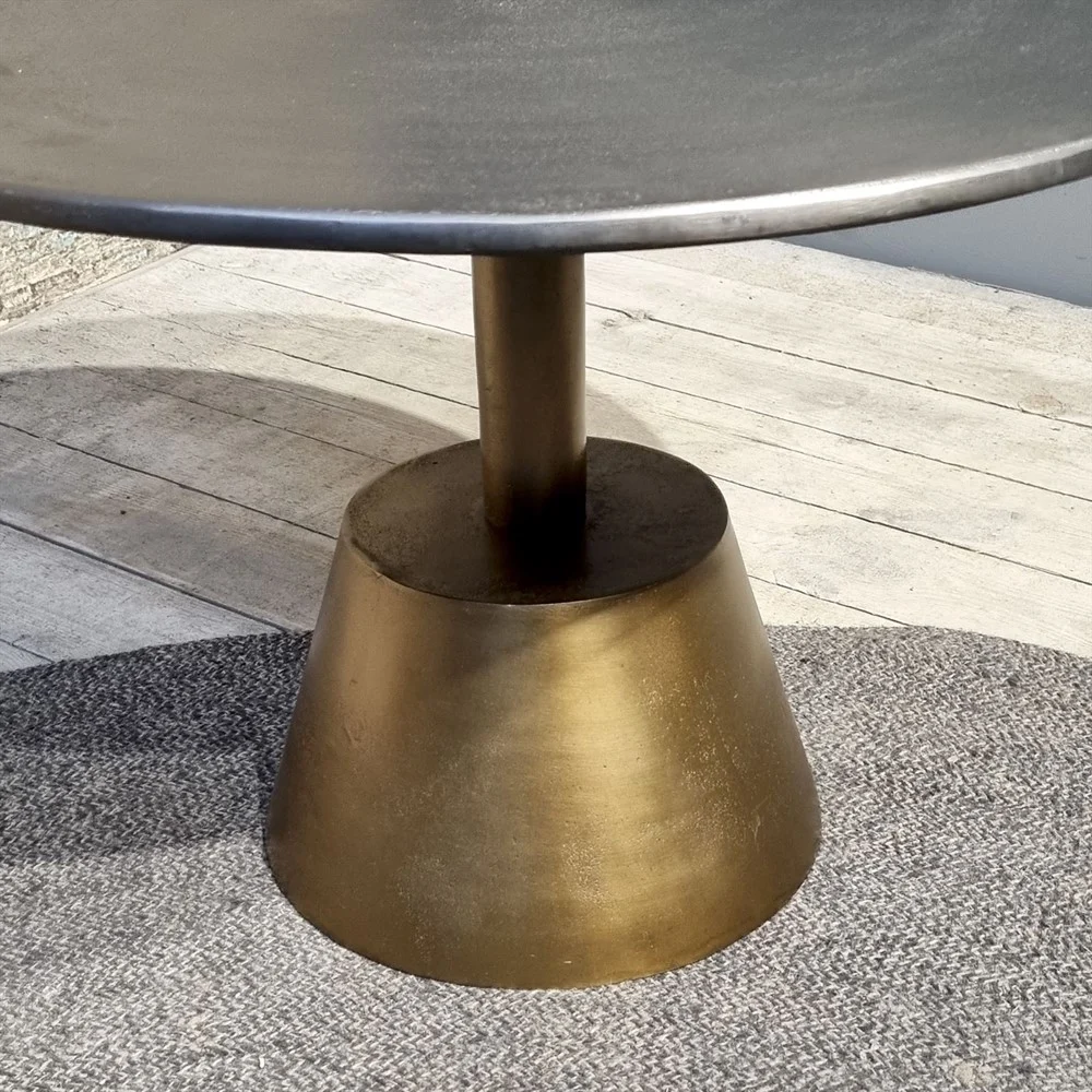 De Sidetable Kian Goud Zilver heeft een rond, donker tafelblad met een kegelvormig onderstel in metallic goud, op een grijs vloerkleed met textuur op een lichte houten vloer.