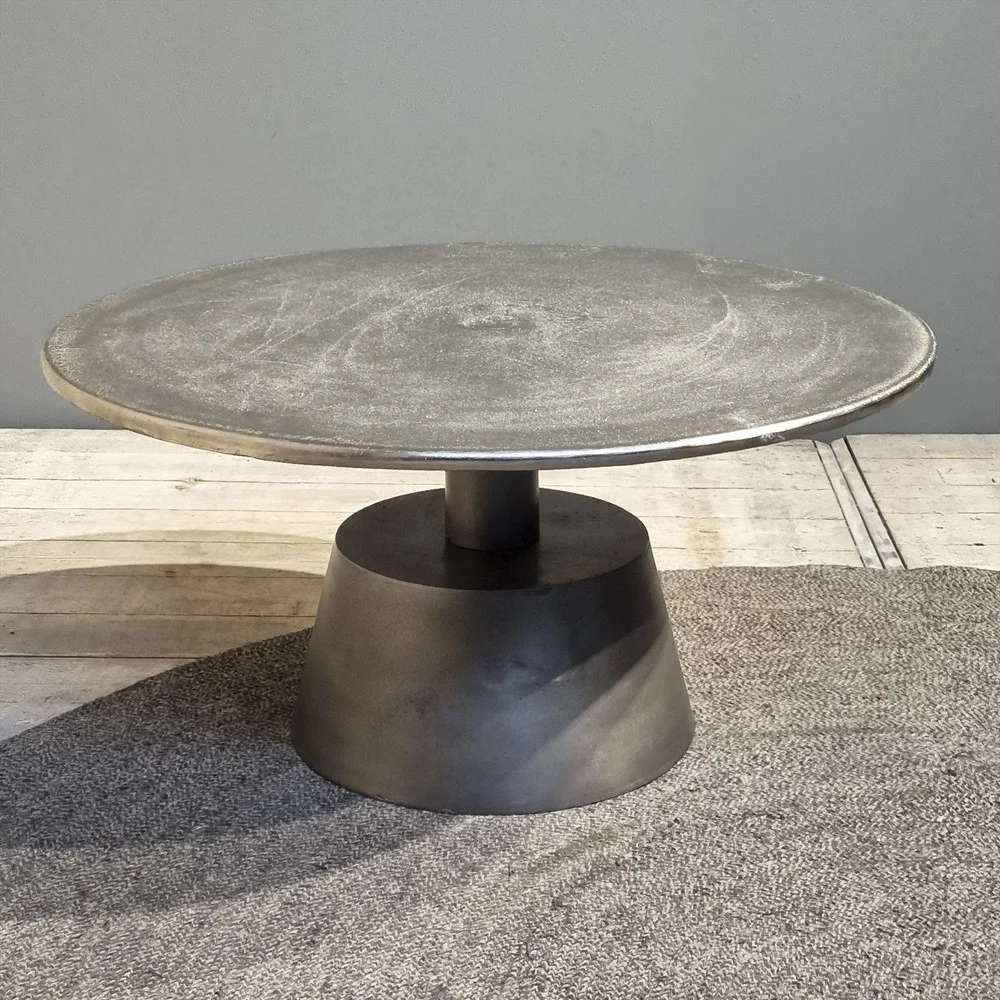 De Salontafel Zilver 82 x 82 CM Vierkant, een moderne vierkante metalen salontafel met een breed plat blad en een stevig onderstel, staat op een grijs vloerkleed tegen een neutrale muur.