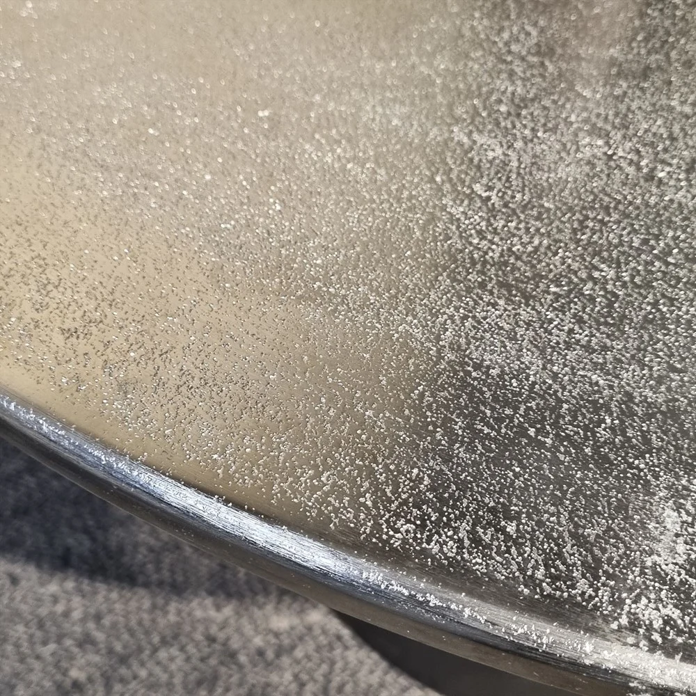 Een close-up van de Salontafel Zilver 82 x 82 CM Vierkant toont het ronde metalen oppervlak met een matte, glinsterende textuur tegen een licht wazige achtergrond.