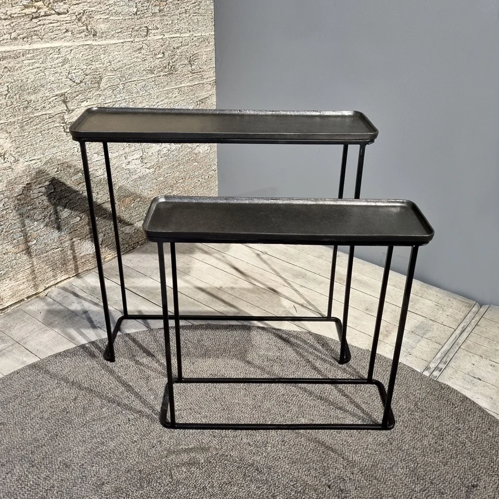 De Sidetable Roan Set van 2 bestaat uit twee zwart metalen consoletafels van verschillende hoogte en grootte, elk met een rechthoekig blad en slanke poten, geplaatst op een grijs vloerkleed tegen een muur met textuur en een lichte houten vloer.