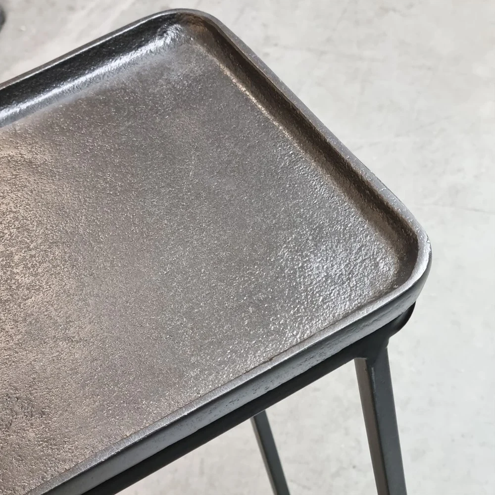 Close-up van de Sidetable Roan Set van 2: vierkante metalen bladen met opstaande randen en gestructureerde oppervlakken, ondersteund door zwarte metalen poten, getoond op een lichtgekleurde vloer.