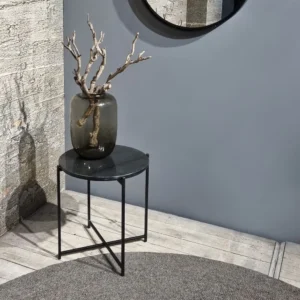 De Sidetable Zwart Marmeren Blad 49" heeft dunne metalen poten en een zwart rond blad. Er staat een vaas in van donker glas met kale takjes, naast gestructureerd beton en grijze muren onder een ronde spiegel.