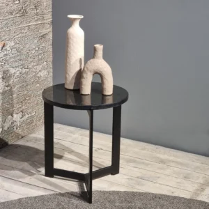 De bijzettafel Onyx, met een rond zwart blad en metalen poten, toont twee crèmekleurige keramieken vazen met verschillende vormen tegen een grijze muur en rustieke houten vloer.