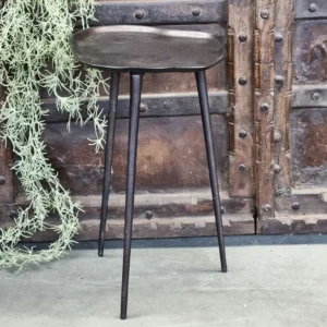 De Sidetable Tamara, een compacte donkerbruine driepotige kruk met een zacht holle zitting, staat op een betonnen vloer naast een groene bladplant en een rustieke houten deur versierd met metalen beslag.