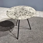 De Sidetable Koper Glas 38" heeft een klein, rond, steenachtig blad met drie dunne zwarte poten en staat op een grijs vloerkleed in een moderne minimalistische kamer.