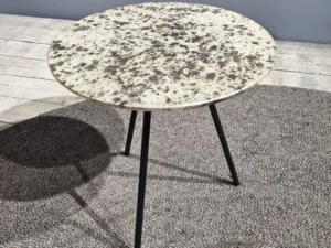 De Sidetable Koper Glas 38" heeft een klein, rond, steenachtig blad met drie dunne zwarte poten en staat op een grijs vloerkleed in een moderne minimalistische kamer.