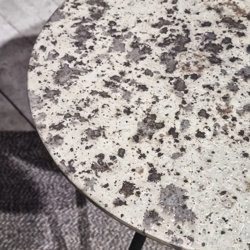 Een close-up van de Sidetable Koper Glas 38" toont het ronde, granietachtige oppervlak in witte, grijze en zwarte tinten, bovenop een tapijt met textuur en patroon.