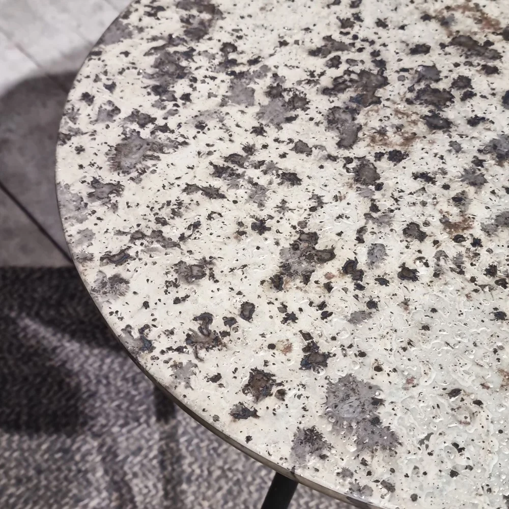 Een close-up van de Sidetable Koper Glas 38" toont het ronde, granietachtige oppervlak in witte, grijze en zwarte tinten, bovenop een tapijt met textuur en patroon.