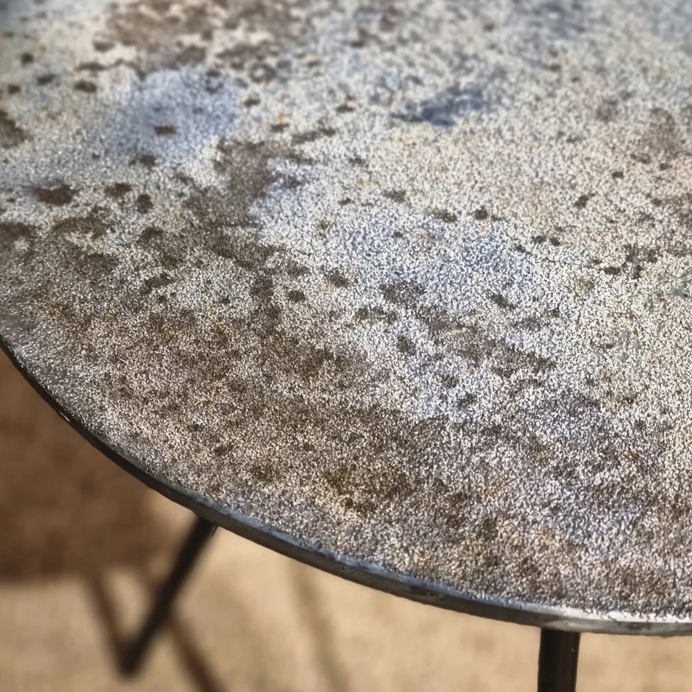 Close-up van de Sidetable Koper Glas 38" toont een rond metalen tafelblad met een verweerd, roestig, gespikkeld oppervlak in bruine, grijze en blauwe tinten. De zwarte rand en een deel van de tafelpoten zijn ook zichtbaar.
