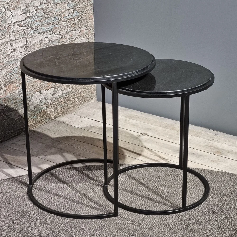 De Sidetable Rond Sienna Set van 2 bestaat uit moderne ronde nesttafels met een zwart metalen frame en donker blad. De kleinere tafel is gedeeltelijk weggestopt onder de grotere, geplaatst op een gestructureerde vloer in de buurt van een stenen en grijze muur.