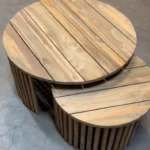Twee Salontafel Teakhout tafels van verschillende afmetingen, met een blad van teakhout met latten, staan naast elkaar op een betonnen vloer, waarbij de kleinere tafel gedeeltelijk onder de grotere tafel is geschoven.