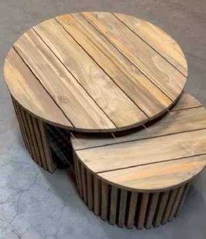 Twee Salontafel Teakhout tafels van verschillende afmetingen, met een blad van teakhout met latten, staan naast elkaar op een betonnen vloer, waarbij de kleinere tafel gedeeltelijk onder de grotere tafel is geschoven.