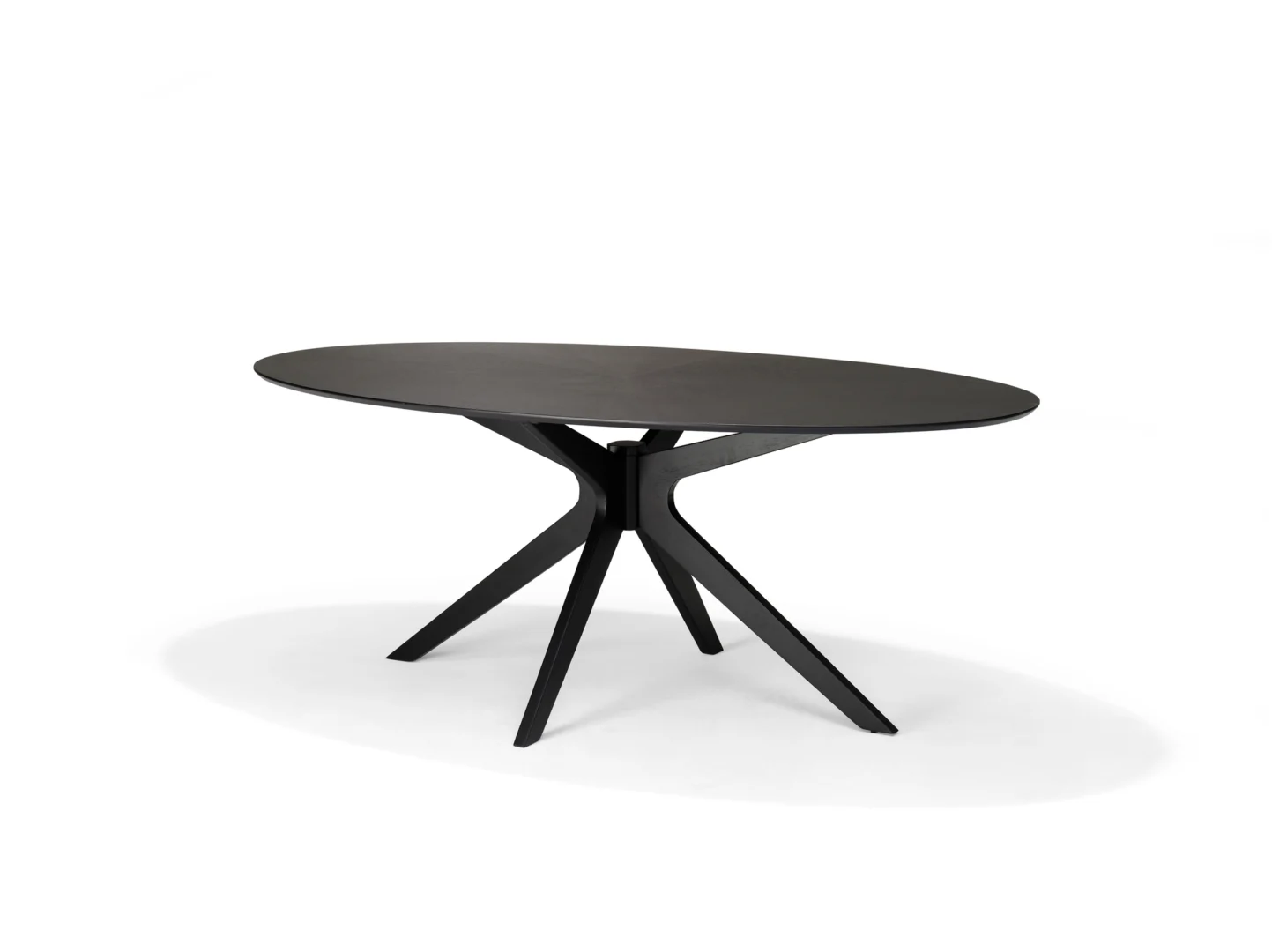 De Julia 200X110 Eettafel Ovaal Zwart Eiken is een moderne, ovaalvormige eettafel van zwart eiken met een glad blad en vier schuine poten die in het midden samenkomen, afgebeeld tegen een effen witte achtergrond.