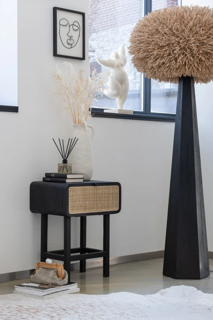 In een moderne, minimalistische kamer staat het Nachtkastje Zwart Industrieel (37x45x60 cm) als een klein zwart tafeltje met boeken, een vaas met gedroogde planten en een rietverdamper. Vlakbij staat een grote decoratieve staande lamp en sculpturaal kunstwerk op de vensterbank.