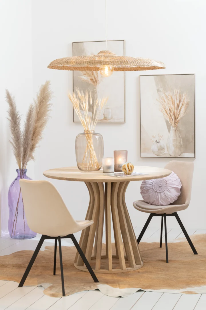 De Tafel Eli Mangohout Natuurlijk is ingericht met twee beige stoelen, een vaas met gedroogd gras, kaarsen, pampagrasdecor, neutrale kunstwerken, een geweven hanglamp en een vloerkleed voor een minimalistische, modern-boho eethoek.