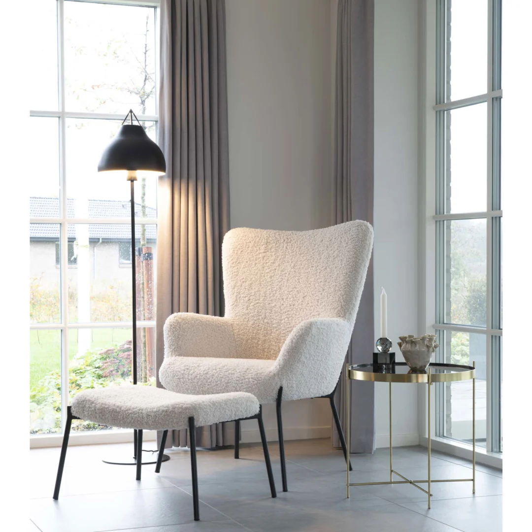 Een gezellige witte fauteuil met een bijpassende voetenbank staat bij grote ramen, naast de Salontafel 48 x 48 CM Vierkant Staal versierd met decor en een brandende kaars. Een zwarte staande lamp staat achter de stoel, met lichtgrijze gordijnen die het tafereel omlijsten.