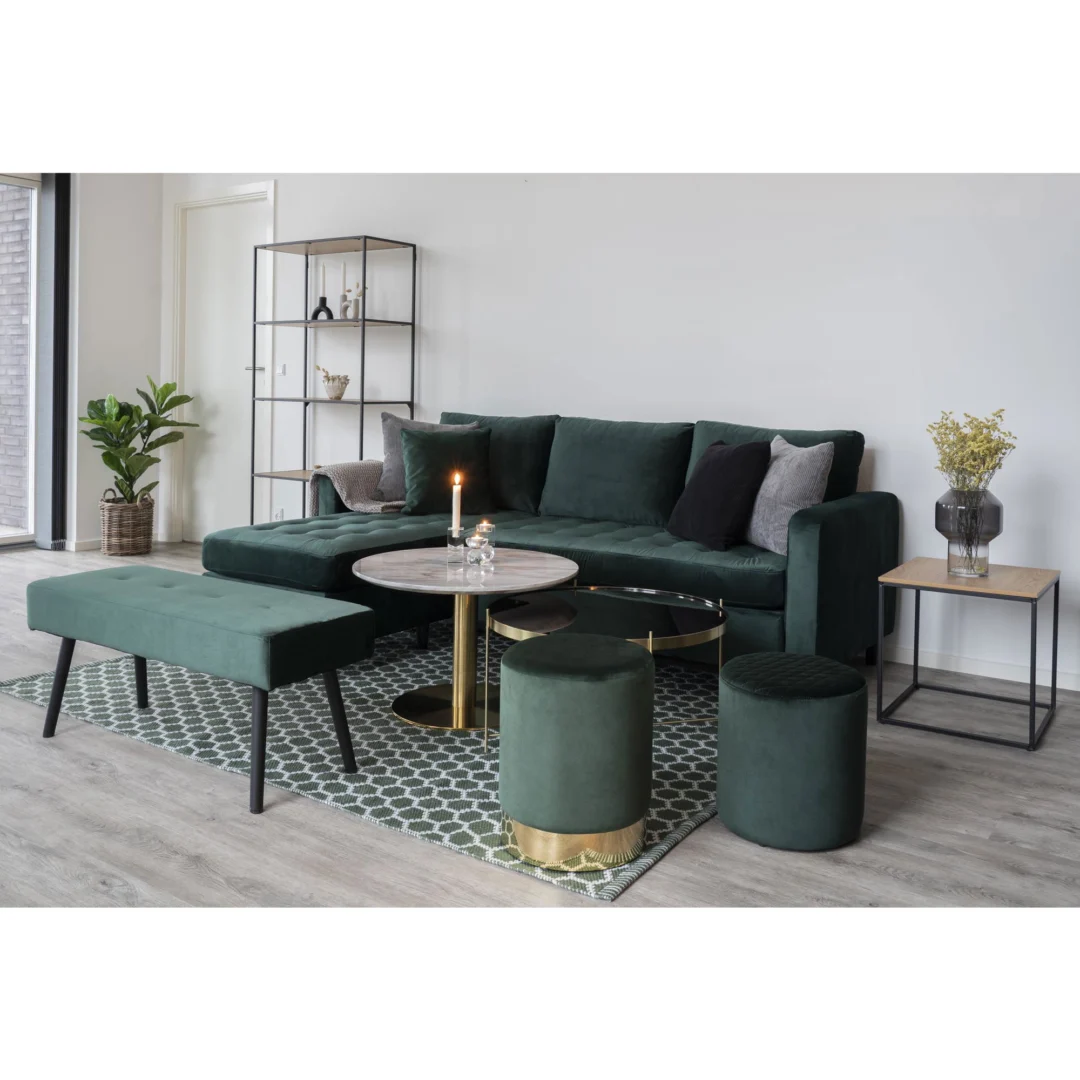 Een moderne woonkamer is voorzien van een groen fluwelen bankstel, bijpassende bank, voetenbank, poef, twee Salontafel Messing, Wit 70 x 45 CM tafels met een vierkant stalen frame, een geometrisch vloerkleed, planten, een kaars en planken met boeken en ornamenten.