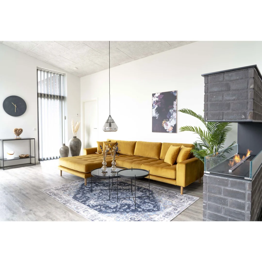 Een moderne woonkamer met een mosterdgele slaapbank, Salontafel Zwart 70 x 40 CM Rond Staal, een blauw vloerkleed met patroon, muurkunst, een open haard met glas, een grote vaas, een kamerplant, een wandklok en hoge ramen die veel licht binnenlaten.