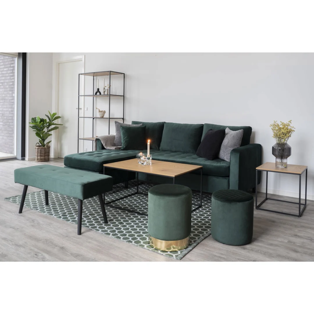 Een moderne woonkamer met een donkergroene fluwelen bank, bijpassende bank, twee ronde voetenbanken, geometrisch vloerkleed, metalen plank, potplant, gele bloemen in een vaas en een Salontafel 60 x 45 CM Rechthoek van spaanplaat als salontafel.