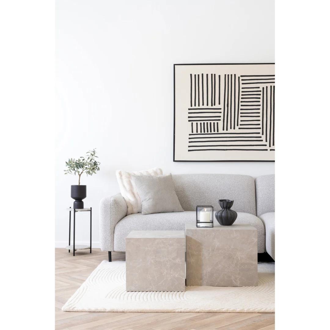 Een moderne woonkamer met een lichtgrijze bank, salontafels Vierkant Marmer 40 x 36 CM, een crèmekleurig vloerkleed, een zwarte vaas, een kaars, een zwart bijzettafeltje met een plant en abstracte zwart-witte muurkunst. De inrichting is minimaal en neutraal.
