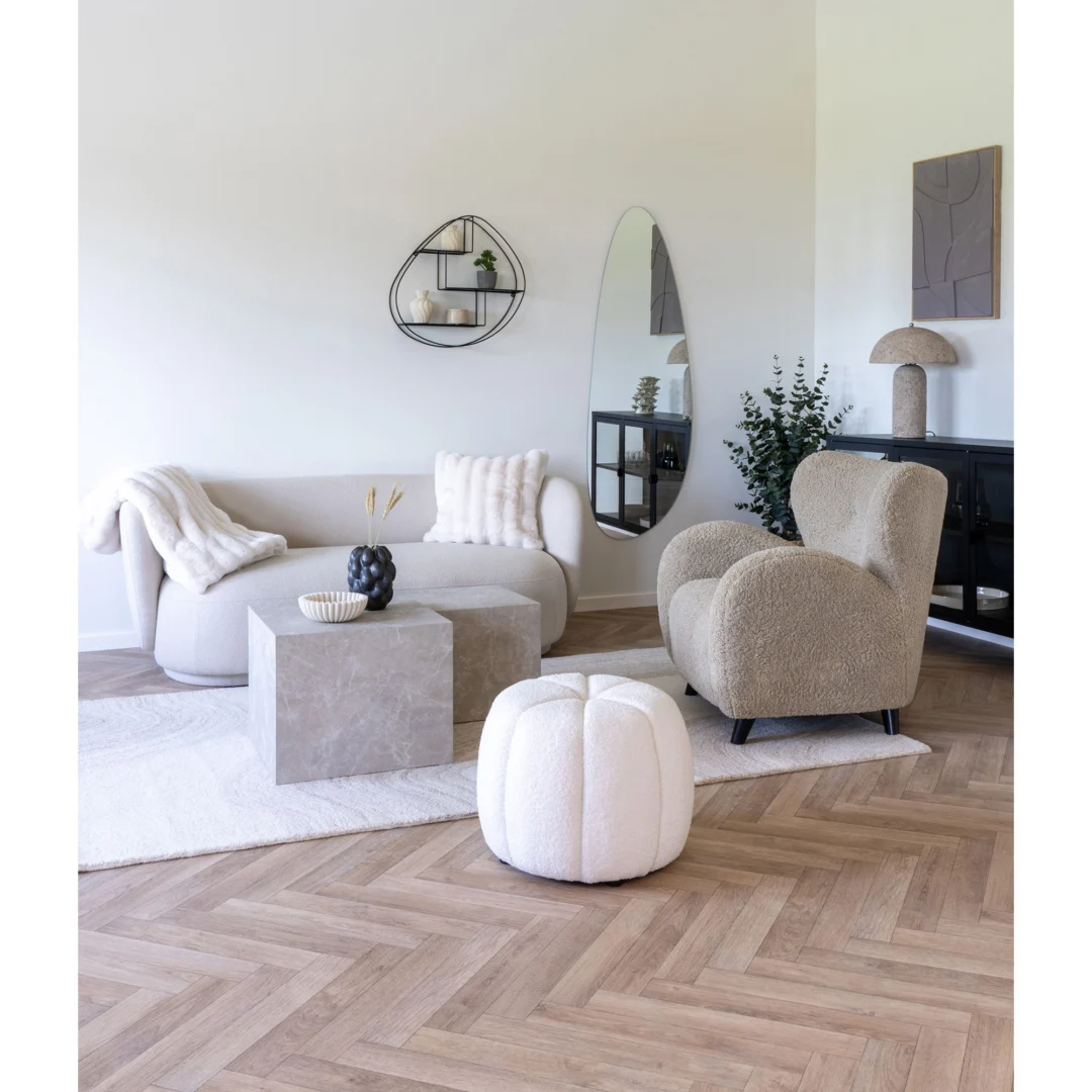 Een moderne woonkamer met een gebogen beige bank, een fauteuil met structuur, Salontafel 40 x 36 CM Vierkant Marmer als salontafel, een witte poef en een minimalistische inrichting. Een grote spiegel en planten verheffen de luchtige ruimte met visgraat houten vloeren.