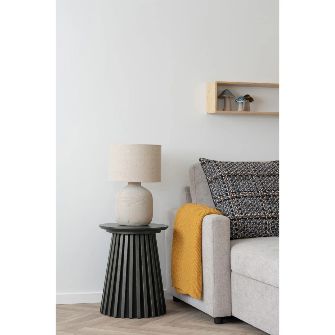 Een moderne woonkamer heeft een lichtgrijze bank met een kussen met patroon en een gele plaid. Naast de bank staat de Osaka Bijzettafel, een ronde zwarte bijzettafel met daarop een beige lamp. Een kleine houten plank met decor hangt aan de witte muur.