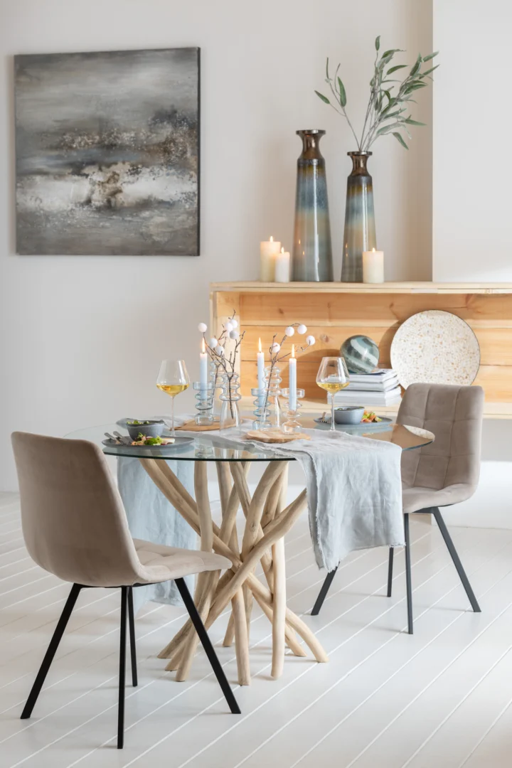 Een moderne eethoek met de Eettafel Takken Rond Teakhout Natuurlijk/Glas voor twee, met beige stoelen, blauwgrijs linnen, kaarsen, wijnglazen en een stijlvolle inrichting. Een schilderij, vazen en kaarsen staan op een houten plank erachter.
