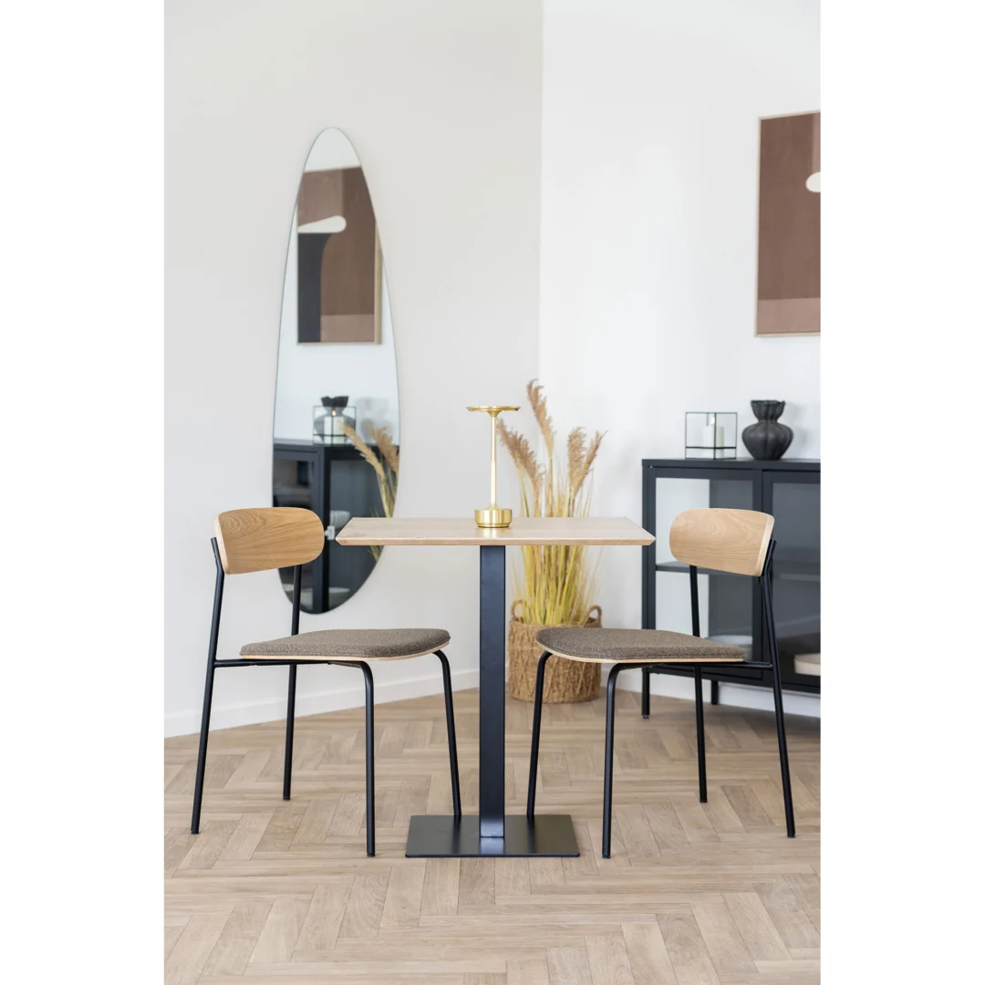 Een lichte, minimalistische eethoek met de Fano Cafétafel, twee stoelen met houten rugleuning en donkere frames, een gouden kandelaar als middenstuk, een hoge ovalen spiegel, pampagras in een vaas en een modern decor.
