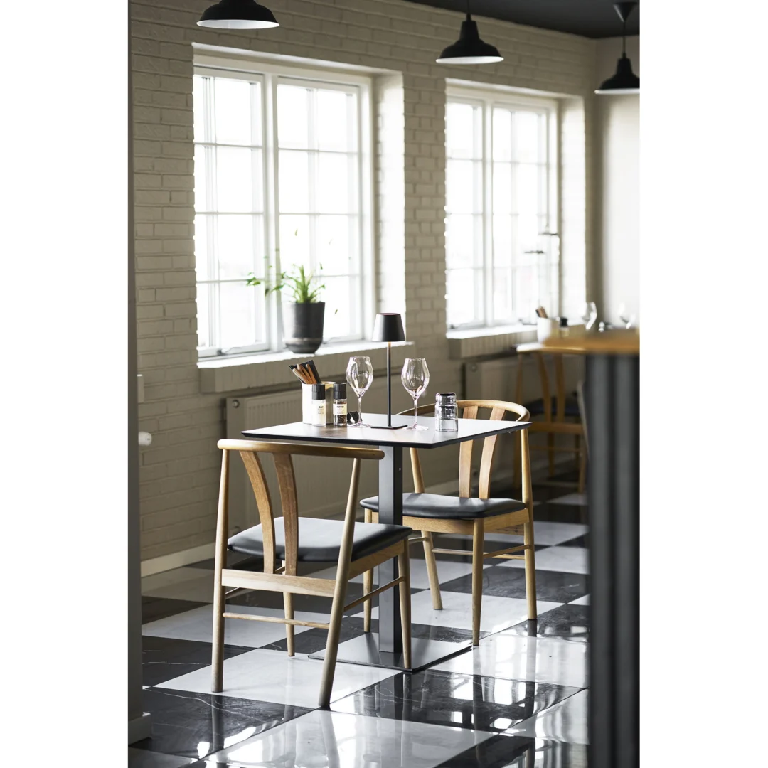 De Fano Cafétafel is ingericht met wijnglazen, bestek en een plant in potten, vergezeld van twee houten stoelen naast grote ramen in een lichte kamer met zwart-wit geblokte vloertegels.