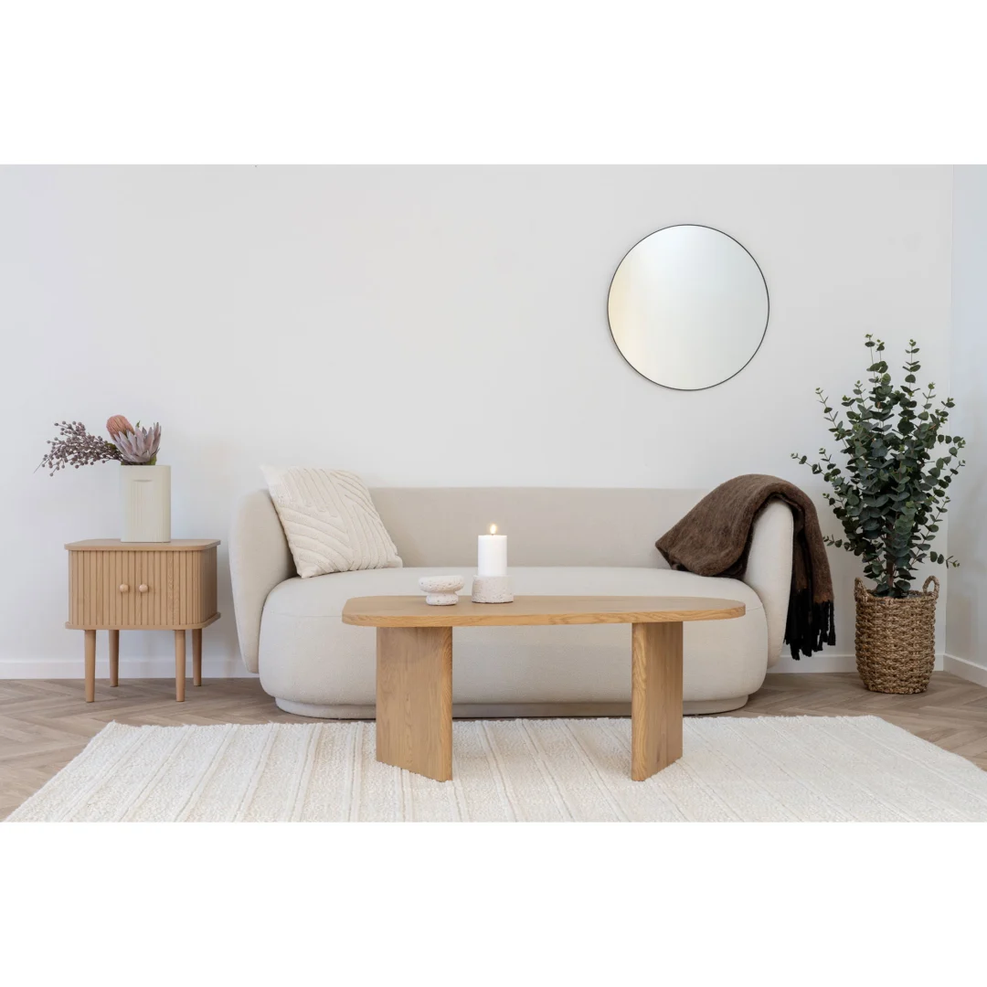 Een minimalistische woonkamer met een lichtbeige bank, een houten salontafel, een brandende kaars, een ronde spiegel op een witte muur, een Nikko Bijzettafel met een vaas en een hoge plant in een gevlochten mand op een licht vloerkleed.
