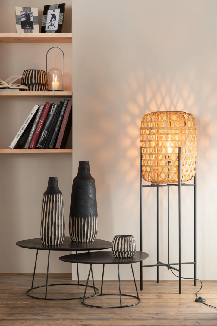 Een gezellige, moderne leefruimte met een geweven vloerlamp die gedessineerd licht werpt, zwarte en beige vazen op de Bijzettafel Bruin 45 x 30 CM Ovaal Metaal en boeken en decor op planken tegen een beige muur.
