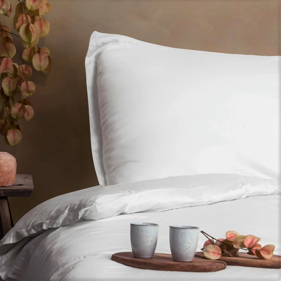 Wit beddengoed met een kussensloop van Tencel. Zachte, neutrale kleuren en natuurlijk licht. Houten dienblad met kopjes en decoratie.
